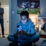 Teen in a bedroom focused on a glowing smartphone while a concerned parent stands in the doorway, with blurred sports poster, headphones, and a fidget toy in the background.